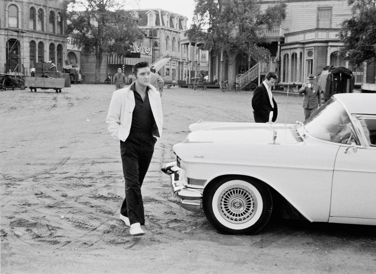 A black and white picture of Elvis walking past a Cadillac.