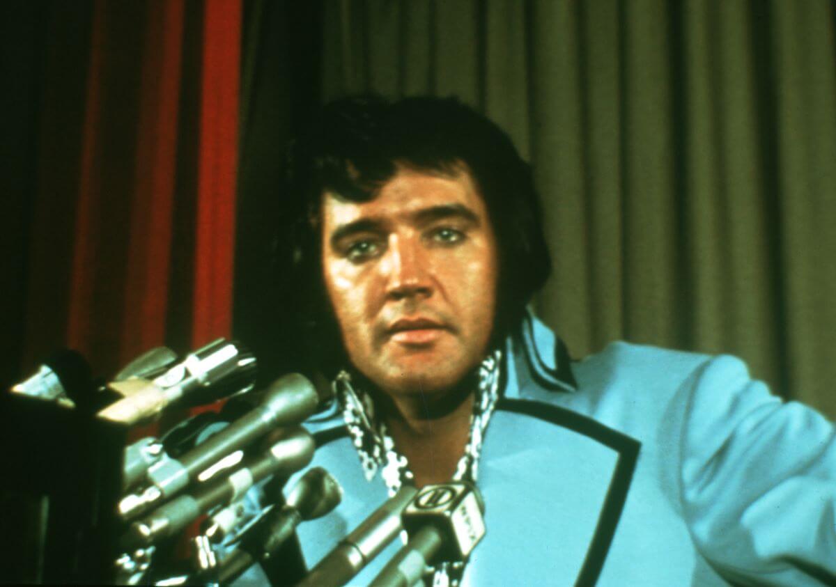 Elvis Presley wears a blue suit and sits in front of multiple microphones.