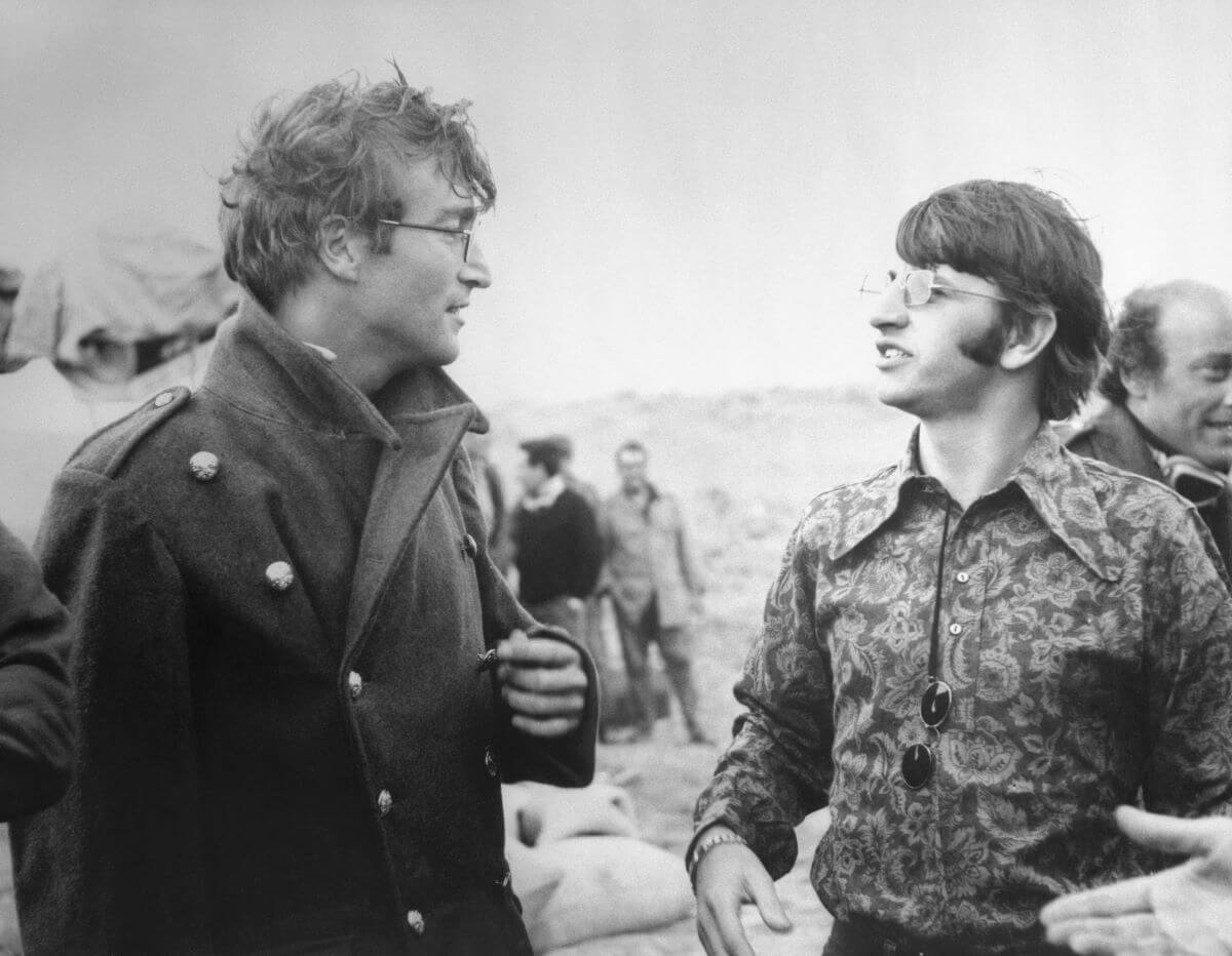 A black and white picture of John Lennon and Ringo Starr in conversation outdoors.