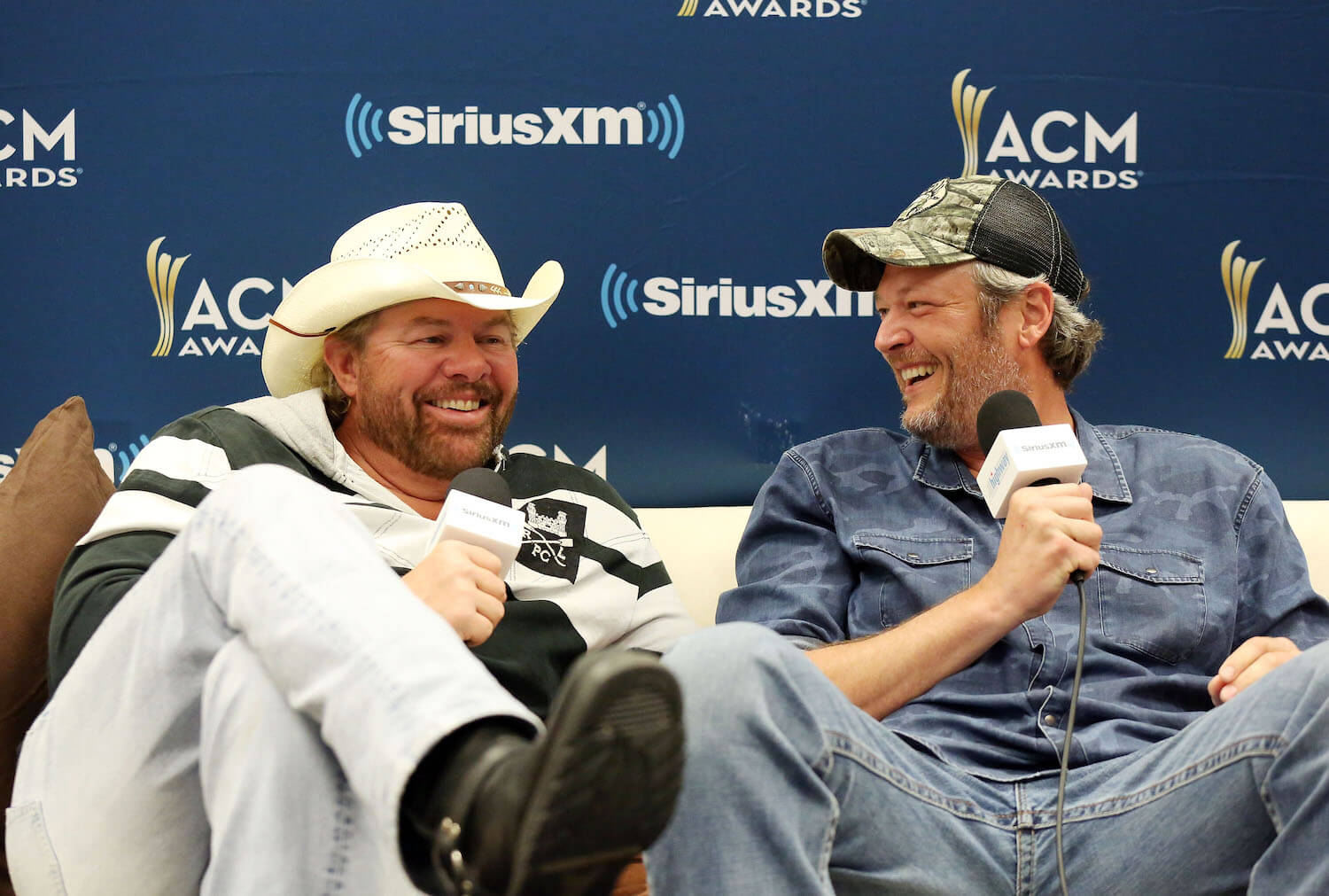 Toby Keith and Blake Shelton sitting on a couch and laughing during an interview in 2018