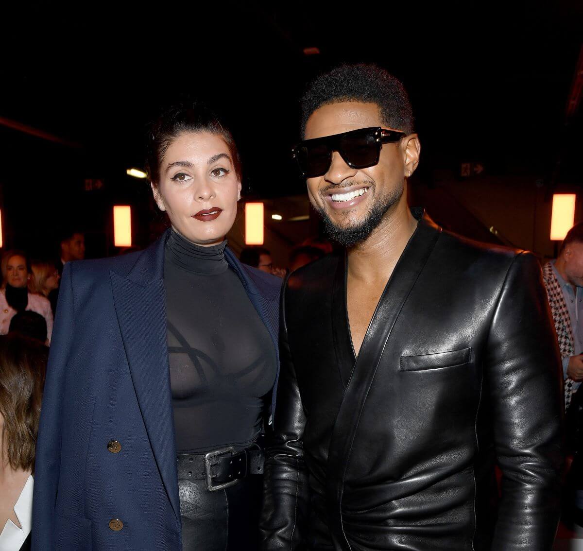 Jenn Goicoechea wears a navy blue blazer and stands next to Usher, who wears a black jacket and sunglasses.