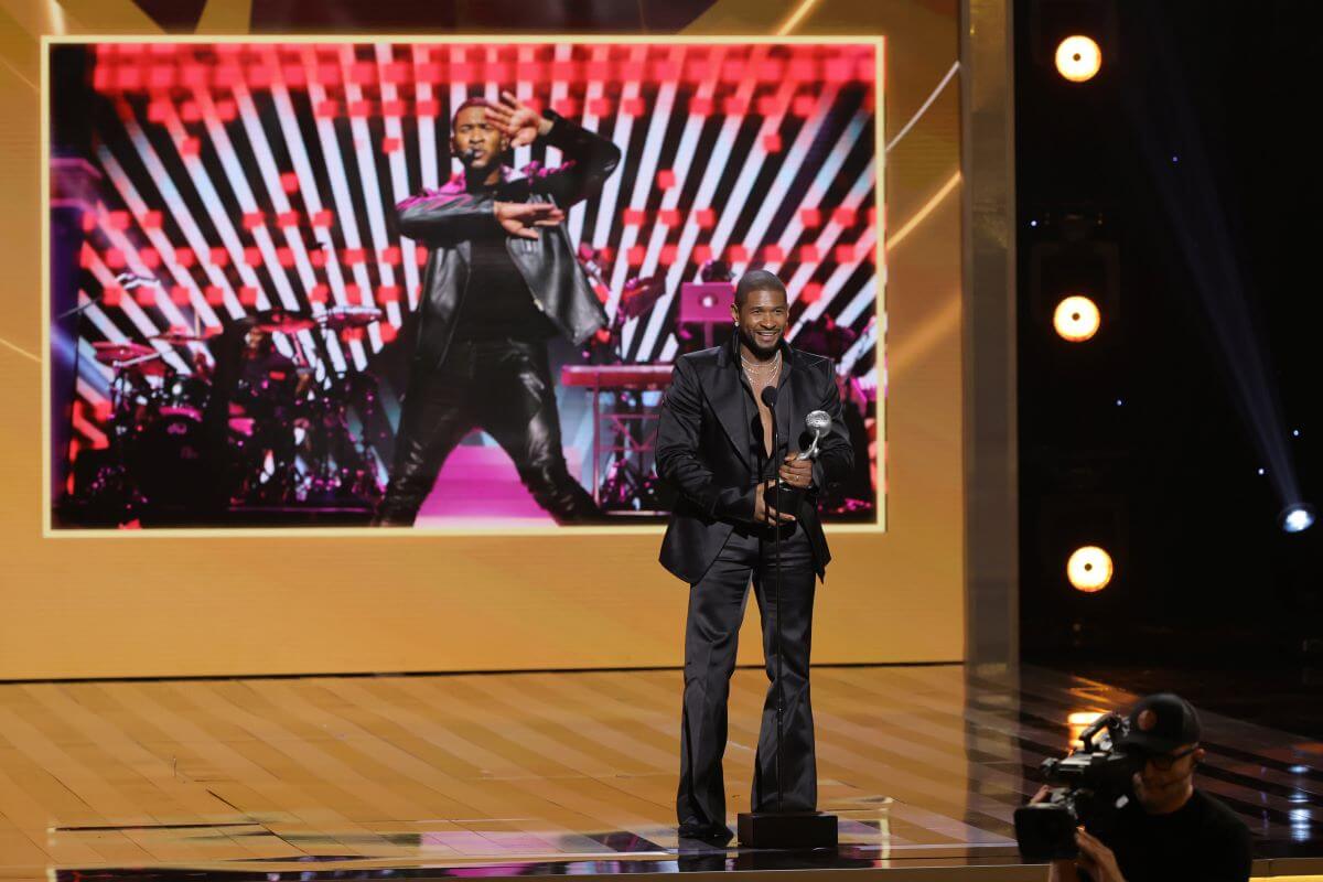 Usher stands in front of a microphone while holding an award. A screen behind him shows him dancing.