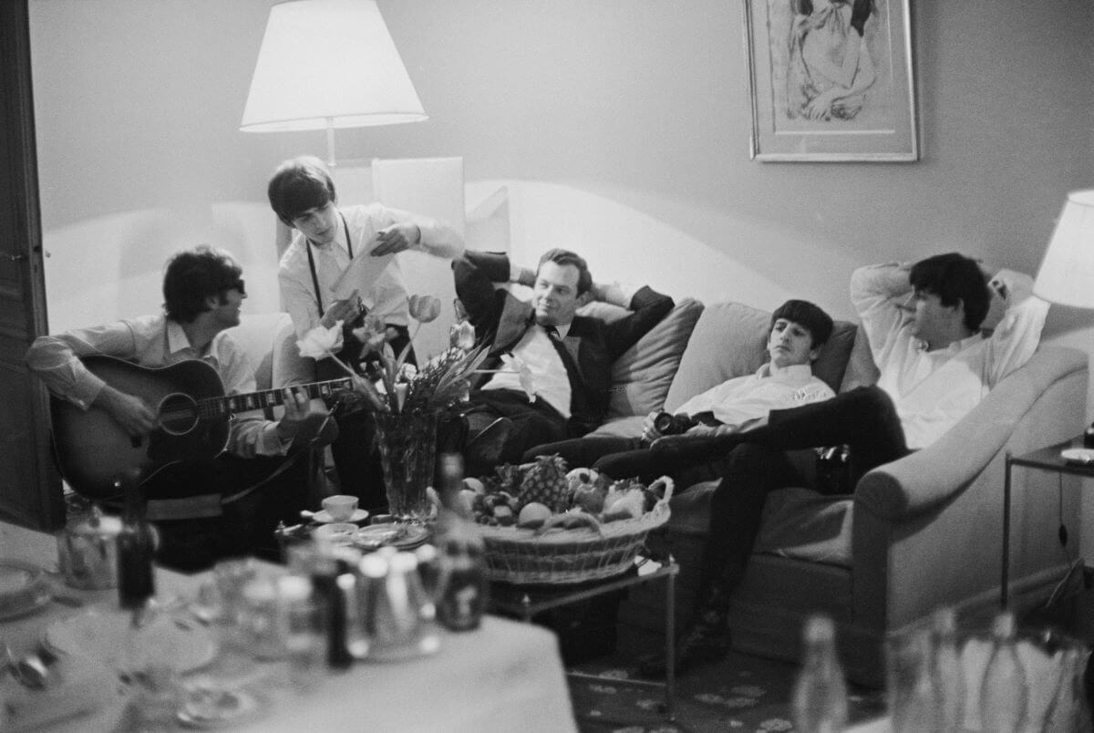 A black and white picture of The Beatles sitting on couches with Brian Epstein.