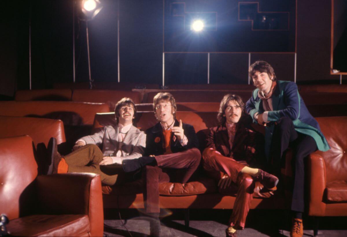 The Beatles sit in a movie theater together. John Lennon points toward the screen.