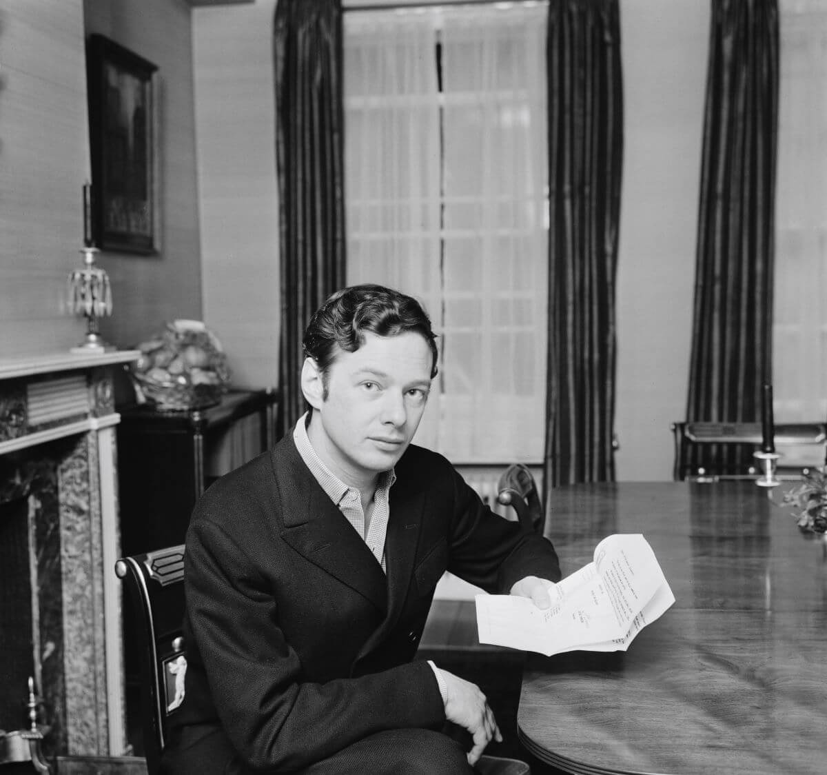 A black and white picture of Brian Epstein sitting at a table with a stack of papers in his hand.