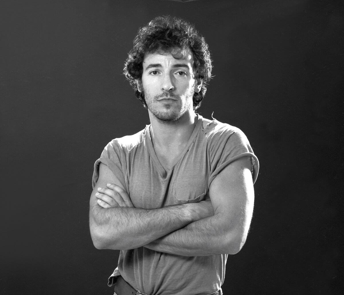 A black and white picture of Bruce Springsteen standing with his arms folded.