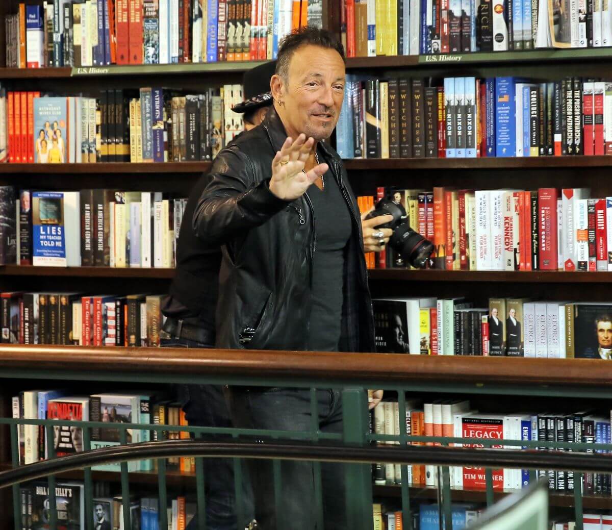 Bruce Springsteen wears black and waves while standing in front of book shelves.