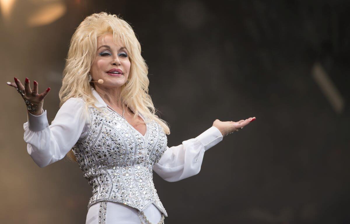 Dolly Parton wears a white, jewel encrusted shirt and lifts her arms in a shrug.