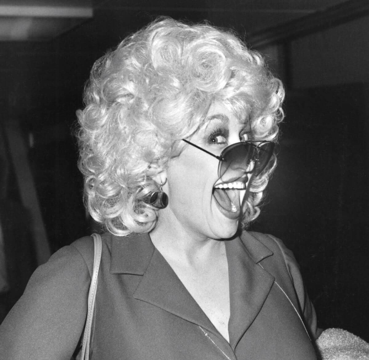 A black and white picture of Dolly Parton wearing sunglasses and looking sideways at the camera. She smiles with her mouth open.