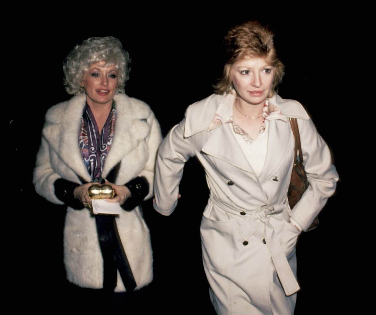 Dolly Parton and Judy Ogle wear coats and walk together. Ogle walks slightly ahead and holds her hand back for Parton.