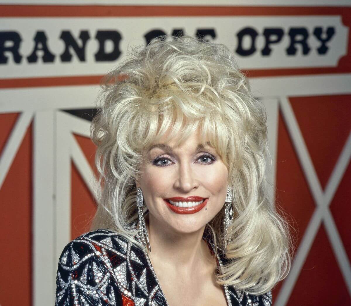 Dolly Parton sits in front of a barn door with a sign that says "Grand Ole Opry."