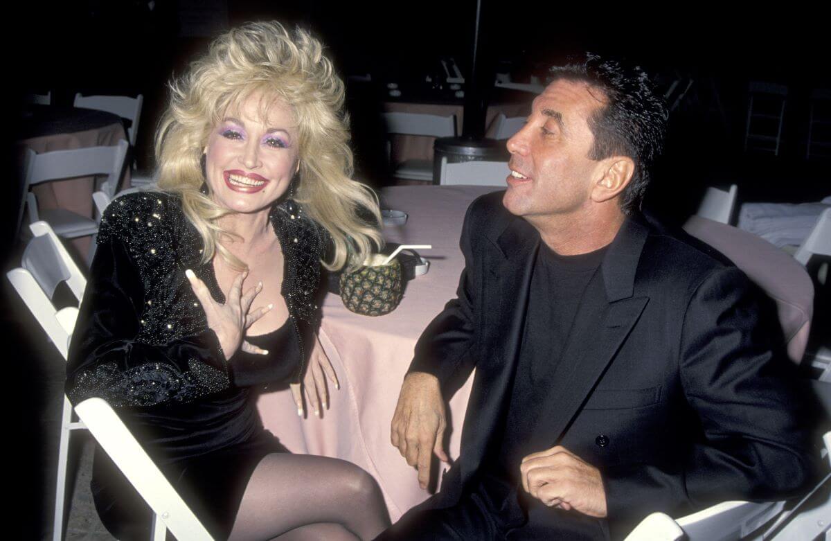 Dolly Parton and Sandy Gallin wear black and sit at a table together.
