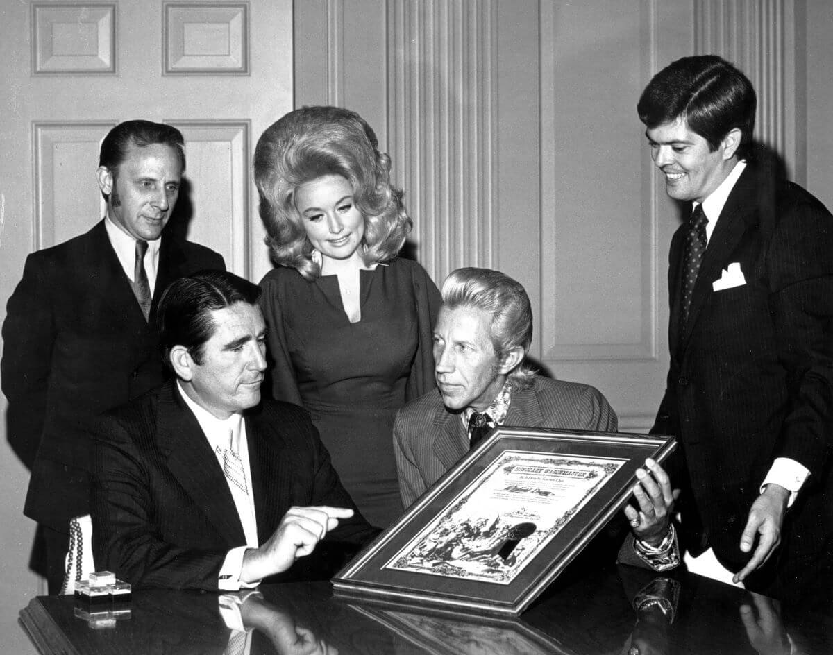 A black and white picture of Dolly Parton and Porter Wagoner standing between three men. Wagoner sits and holds a plaque.