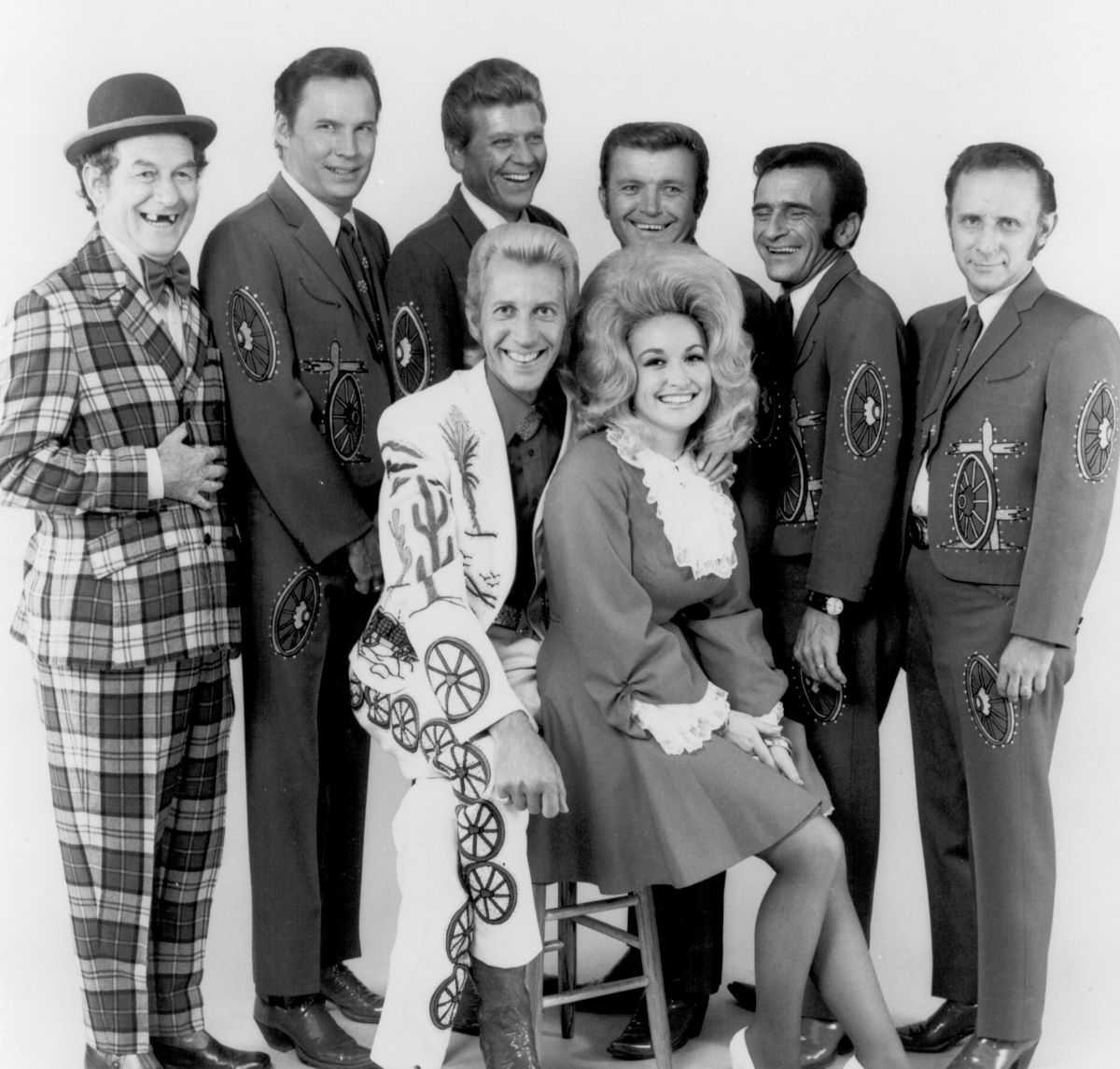 Porter Wagoner and Dolly Parton sit together in front of their backing band of 6 people. Five members of the band wear matching suits and one wears a checkered suit and hat.