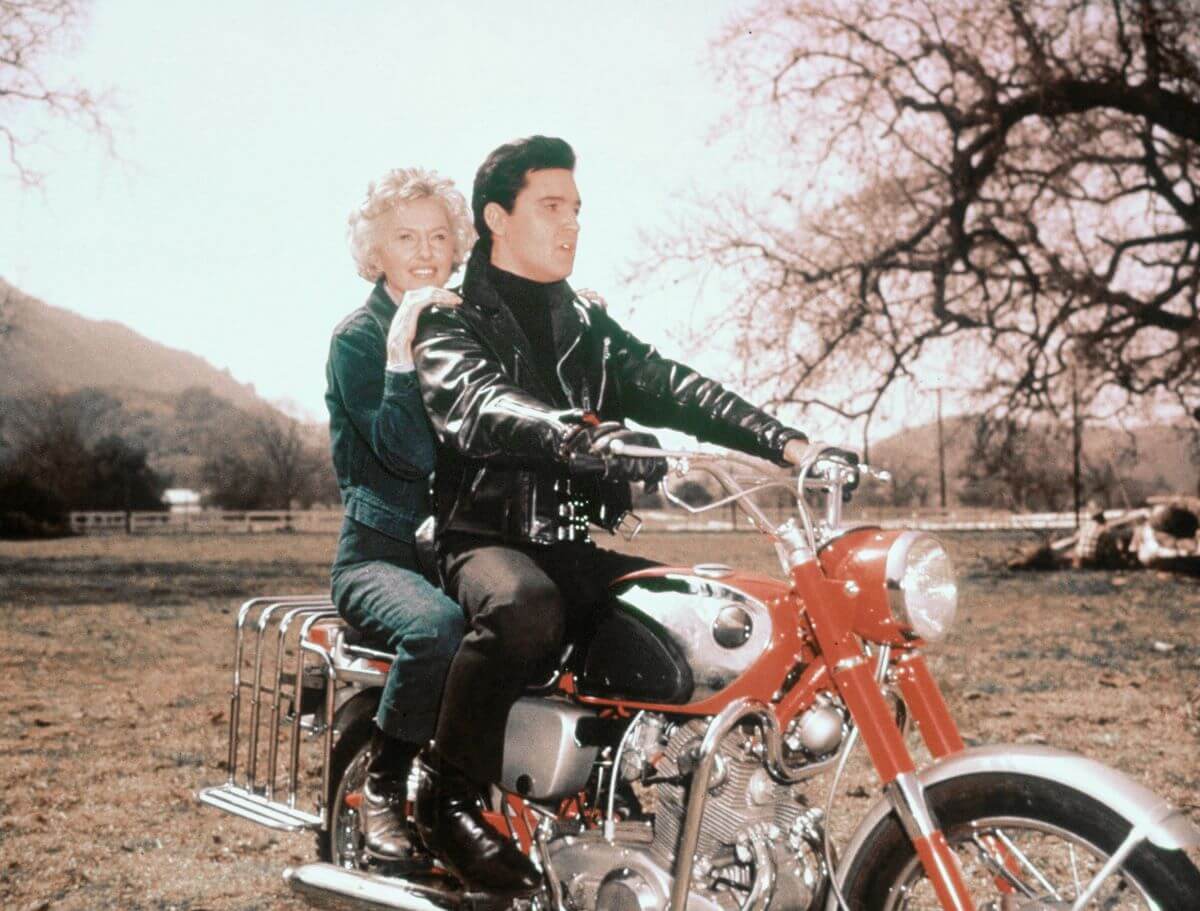 Elvis wears a leather jacket and rides on a red motorcycle with Barbara Stanwyck on the set of 'Roustabout.'