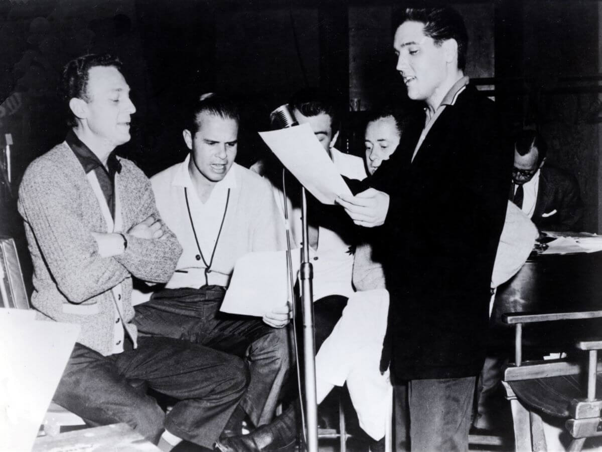 A black and white picture of Elvis holding a piece of paper and singing. The Jordanaires gather around him and sing.