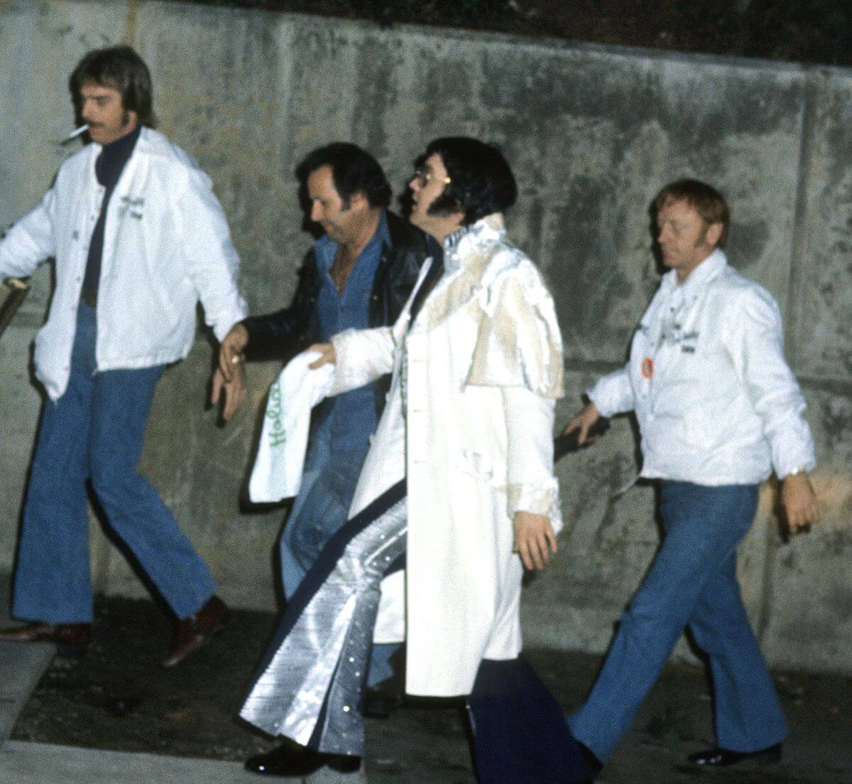 Elvis Presley walks with his bodyguards in front of a concrete wall.