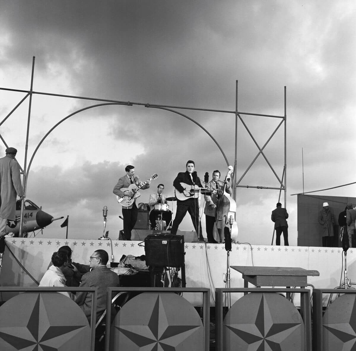 A black and white picture of Elvis Presley performing on a stage with his band.