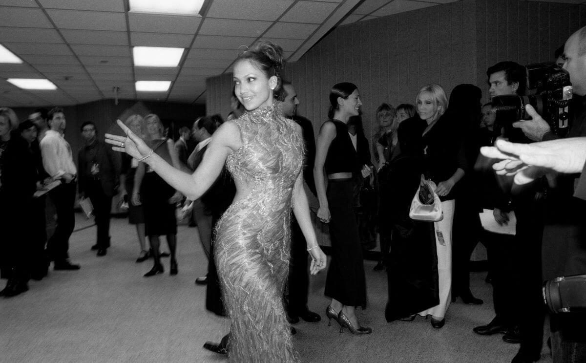 A black and white picture of Jennifer Lopez wearing a dress and walking through a crowded room. She waves at someone behind her.
