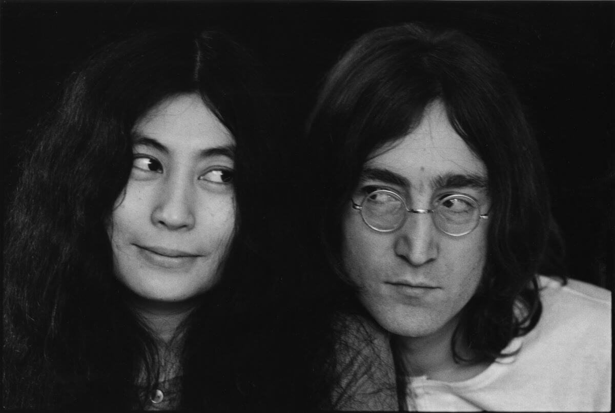 A black and white picture of Yoko Ono and John Lennon looking sideways at each other. He wears glasses.
