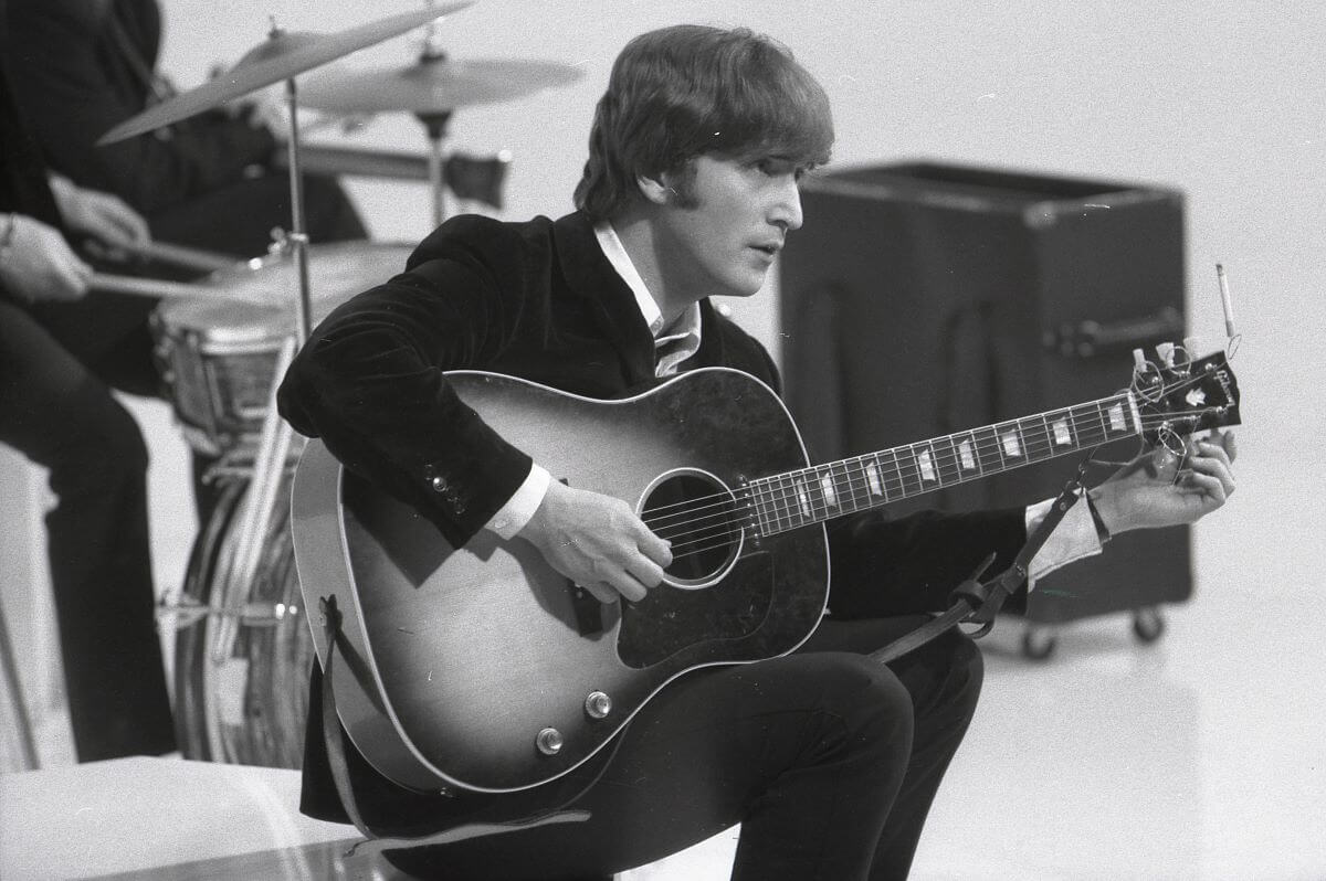 A black and white picture of John Lennon sitting and strumming an acoustic guitar.