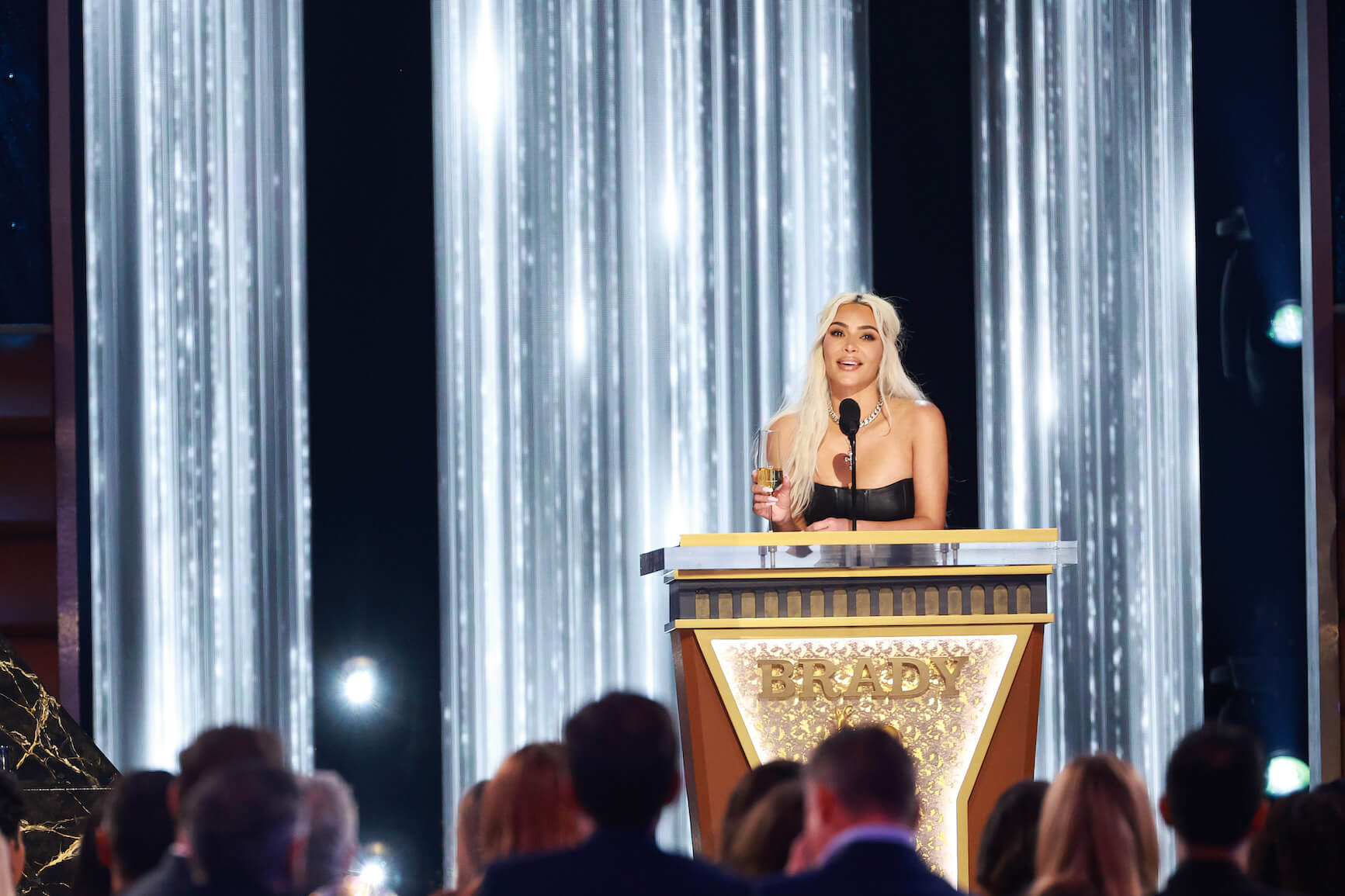 Kim Kardashian standing at a gold podium with the word "Brady" on it in front of a crowd for 'The Roast of Tom Brady'