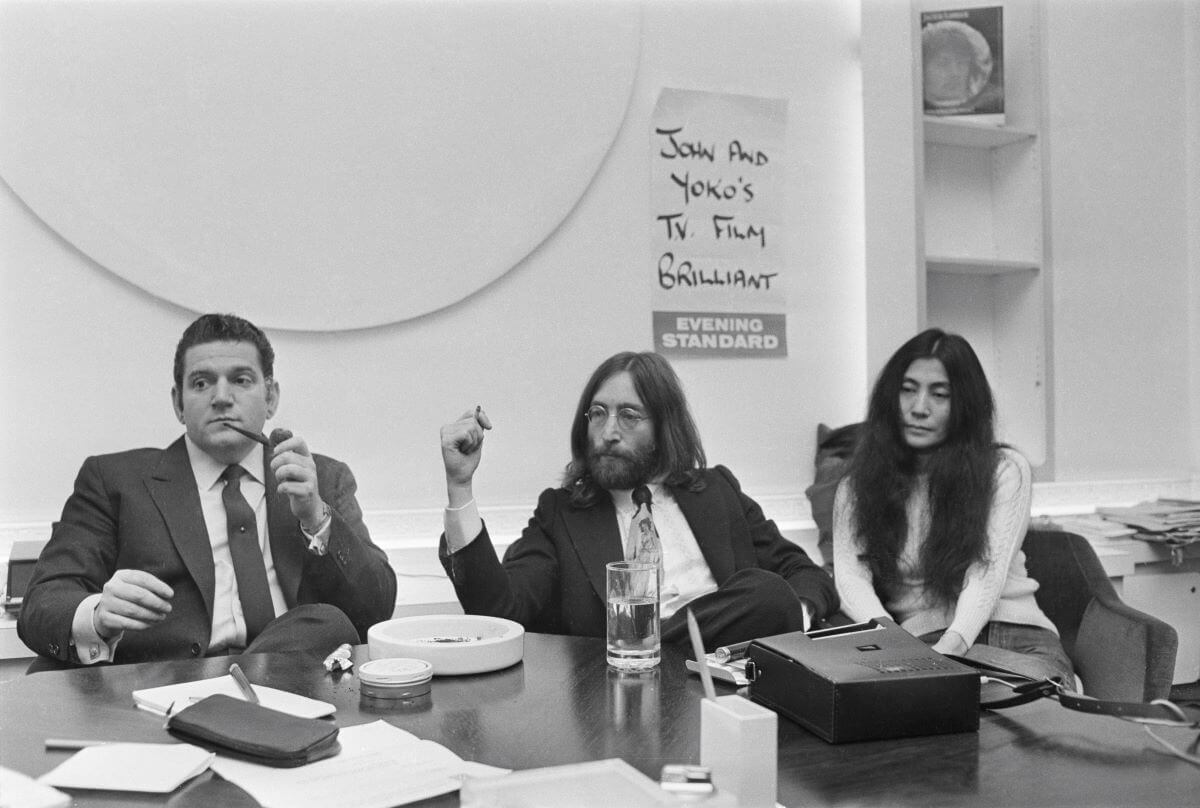 A black and white picture of Allen Klein, John Lennon, and Yoko Ono sitting on one side of a table.