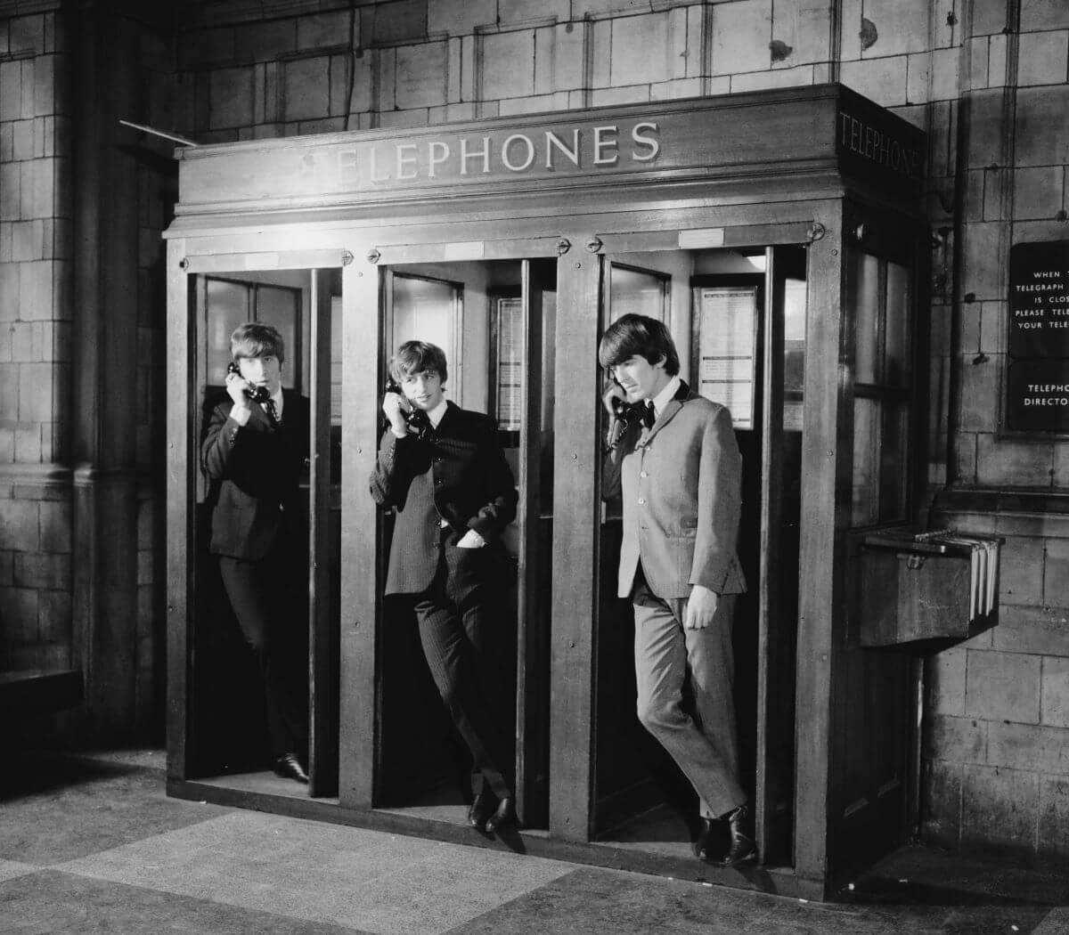 A black and white picture of John Lennon, Ringo Starr, and George Harrison on the phone in telephone booths.