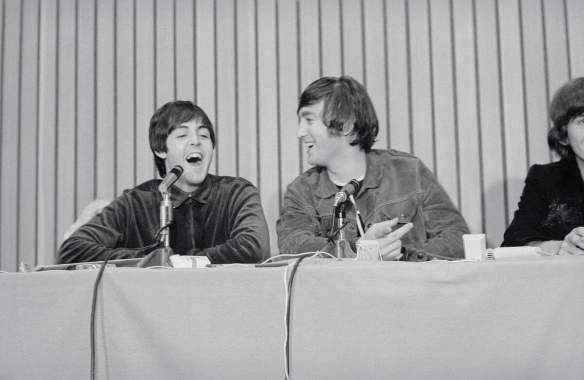 A black and white picture of Paul McCartney and John Lennon sitting with microphones in front of them. They laugh.