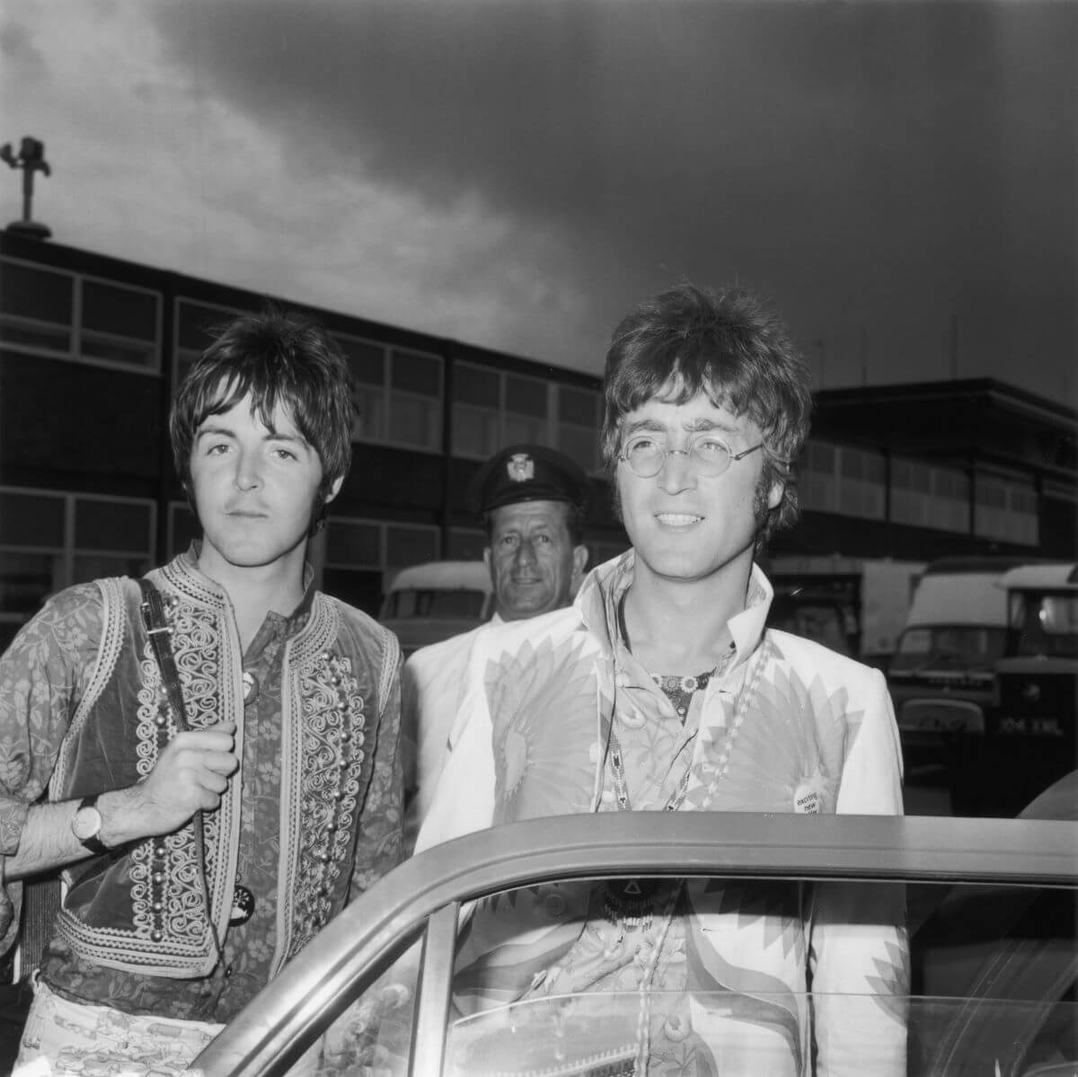 A black and white picture of Paul McCartney and John Lennon standing behind an open car door. A man stands behind them.