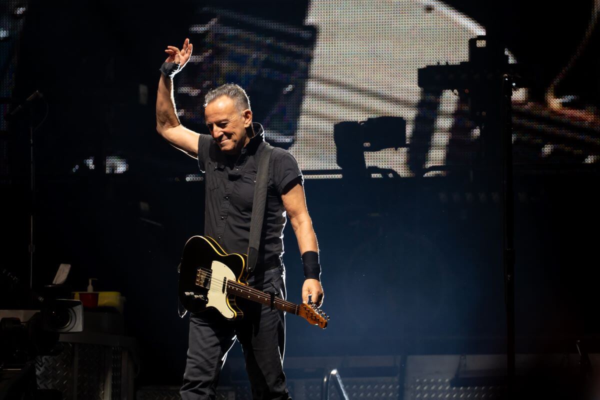 Bruce Springsteen stands on a stage. He lifts one hand in the air and holds a guitar.