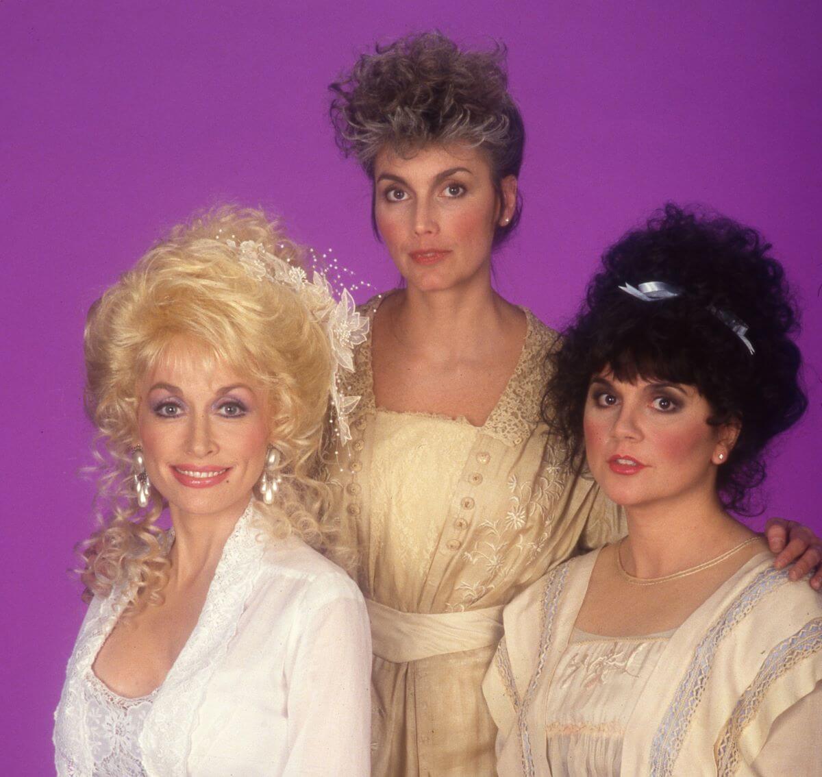Dolly Parton, Emmylou Harris, and Linda Ronstadt pose against a purple background. Harris stands between Parton and Ronstadt, who sit.