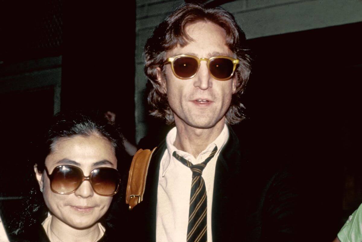 Yoko Ono and John Lennon stand together and wear sunglasses.