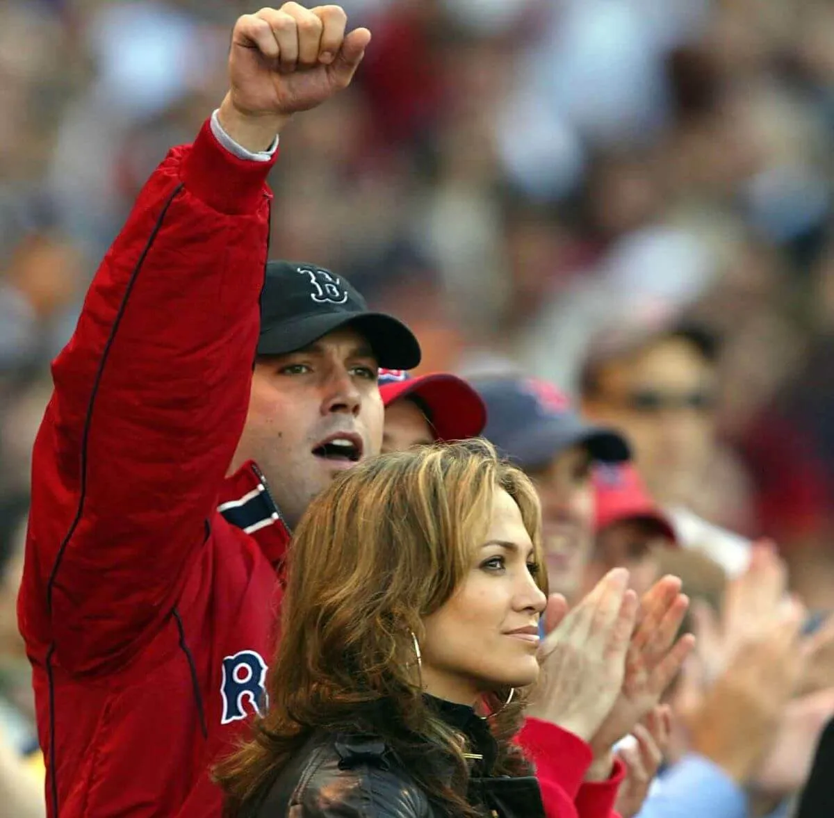 Ben Affleck wears a Red Sox jacket and baseball hat. He lifts his arm into the air at a baseball game. Jennifer Lopez stands next to him wearing a black jacket.