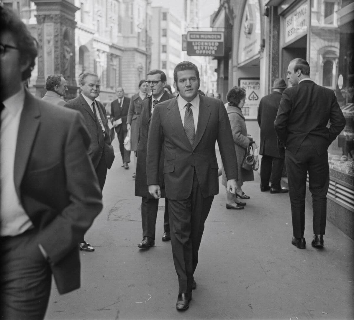 A black and white picture of Allen Klein wearing a suit and walking on the street.
