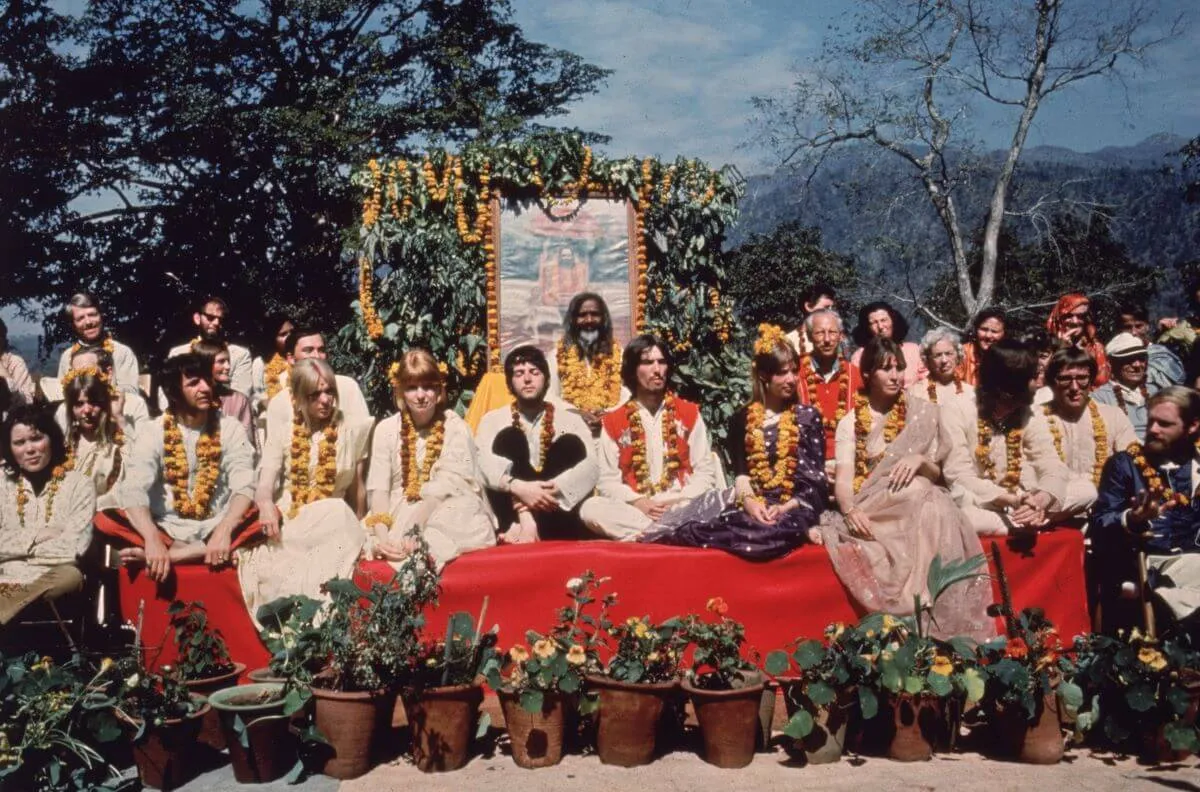 The Beatles sit on a red platform among a group of people while in India.
