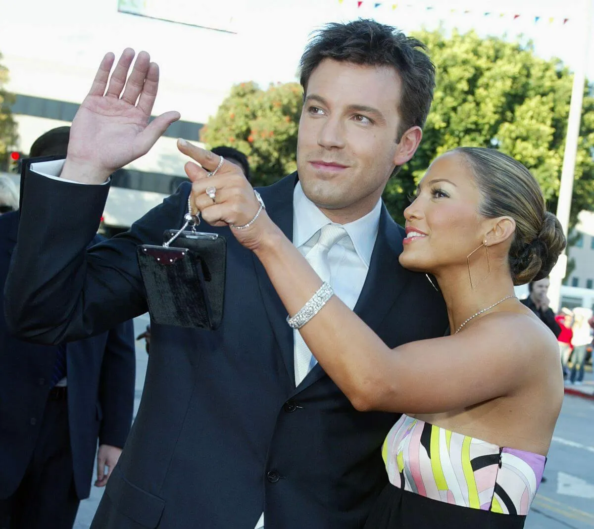 Ben Affleck wears a suit and lifts his arm to wave. Jennifer Lopez stands next to him and points.