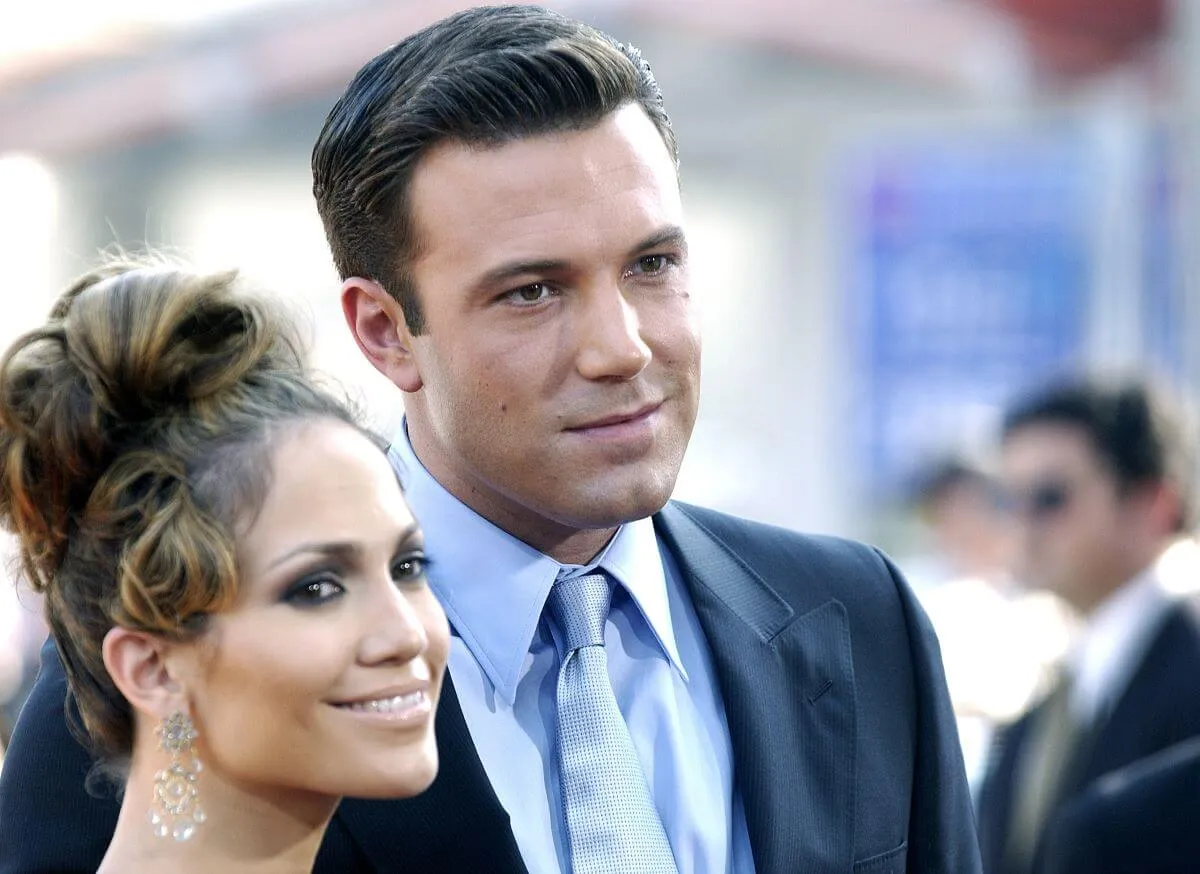 Jennifer Lopez and Ben Affleck stand next to each other. She wears dangling earrings and he wears a suit.