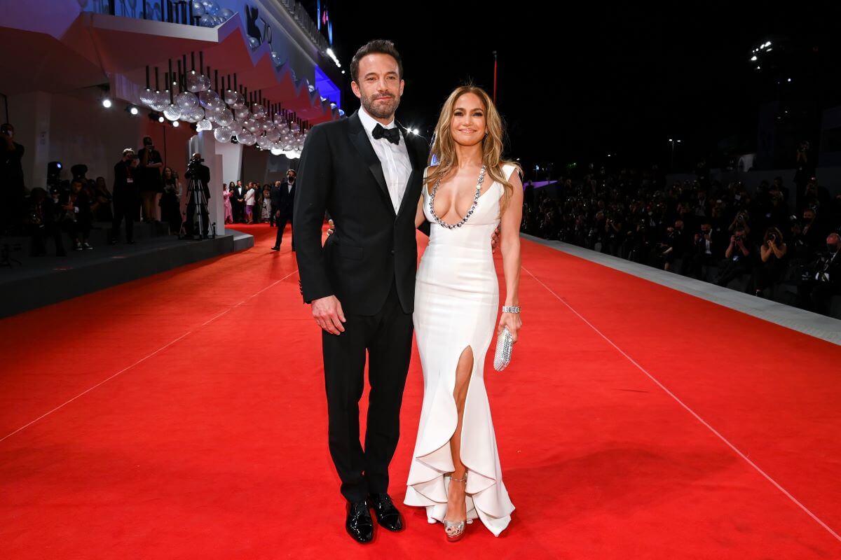 Ben Affleck wears a tuxedo and stands with Jennifer Lopez, who wears a white dress. They stand on a red carpet.