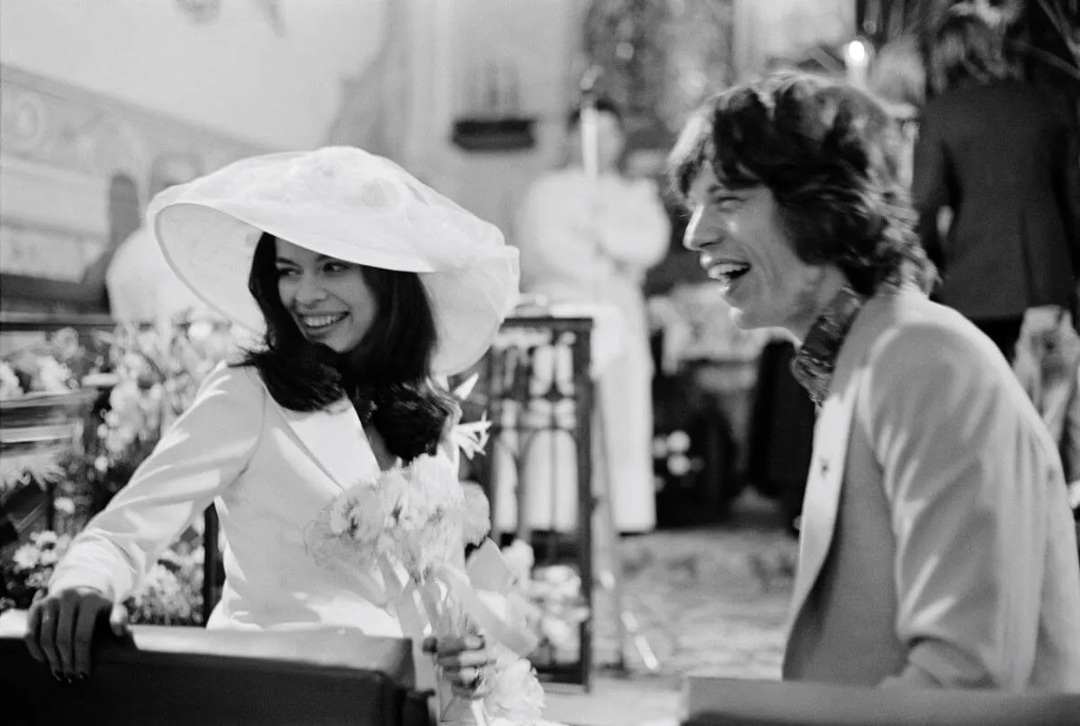A black and white picture of Bianca and Mick Jagger sitting in a church on their wedding day. They both laugh. She wears a hat and holds flowers.