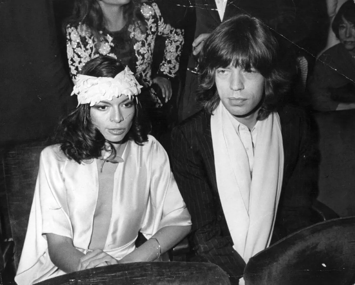 A black and white picture of Bianca Jagger and her husband Mick Jagger sitting together. She wears a headband around her head.