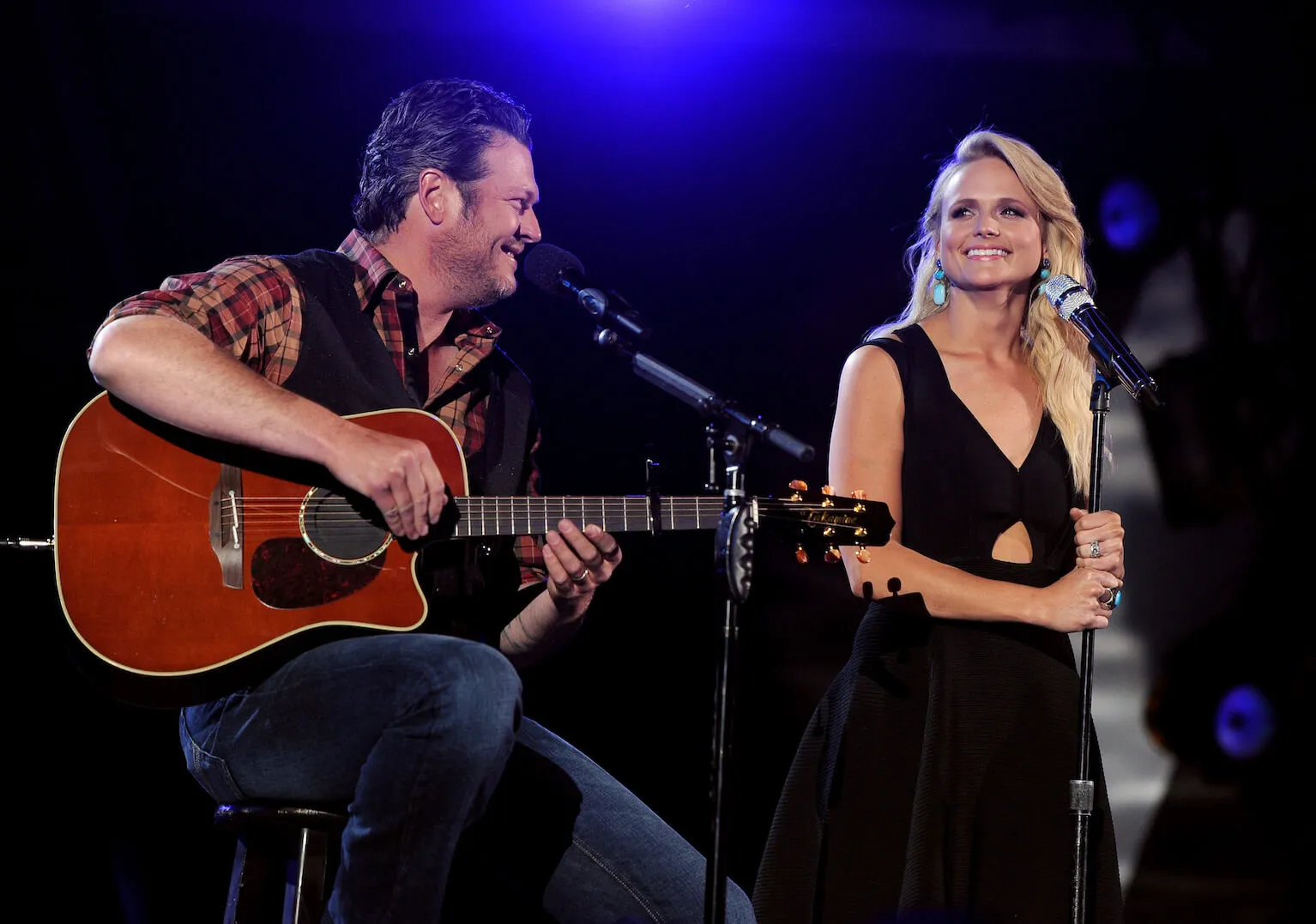 Blake Shelton sitting and holding his guitar on stage next to Miranda Lambert, who's standing in front of a microphone during ACM Presents: An All-Star Salute To The Troops at the MGM Grand Garden Arena in 2014