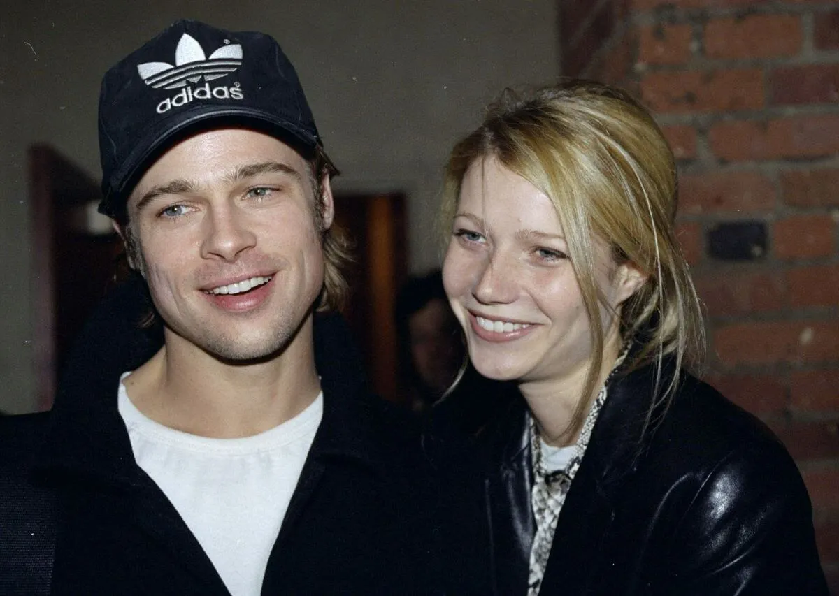 Brad Pitt wears a black Adidas baseball hat and stands next to Gwyneth Paltrow, who wears a black leather jacket.