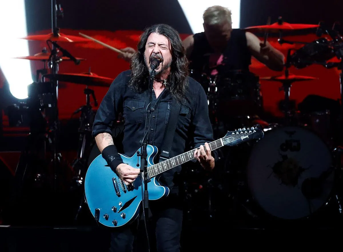 Dave Grohl wears a black shirt and sings into a microphone. He plays a blue guitar.