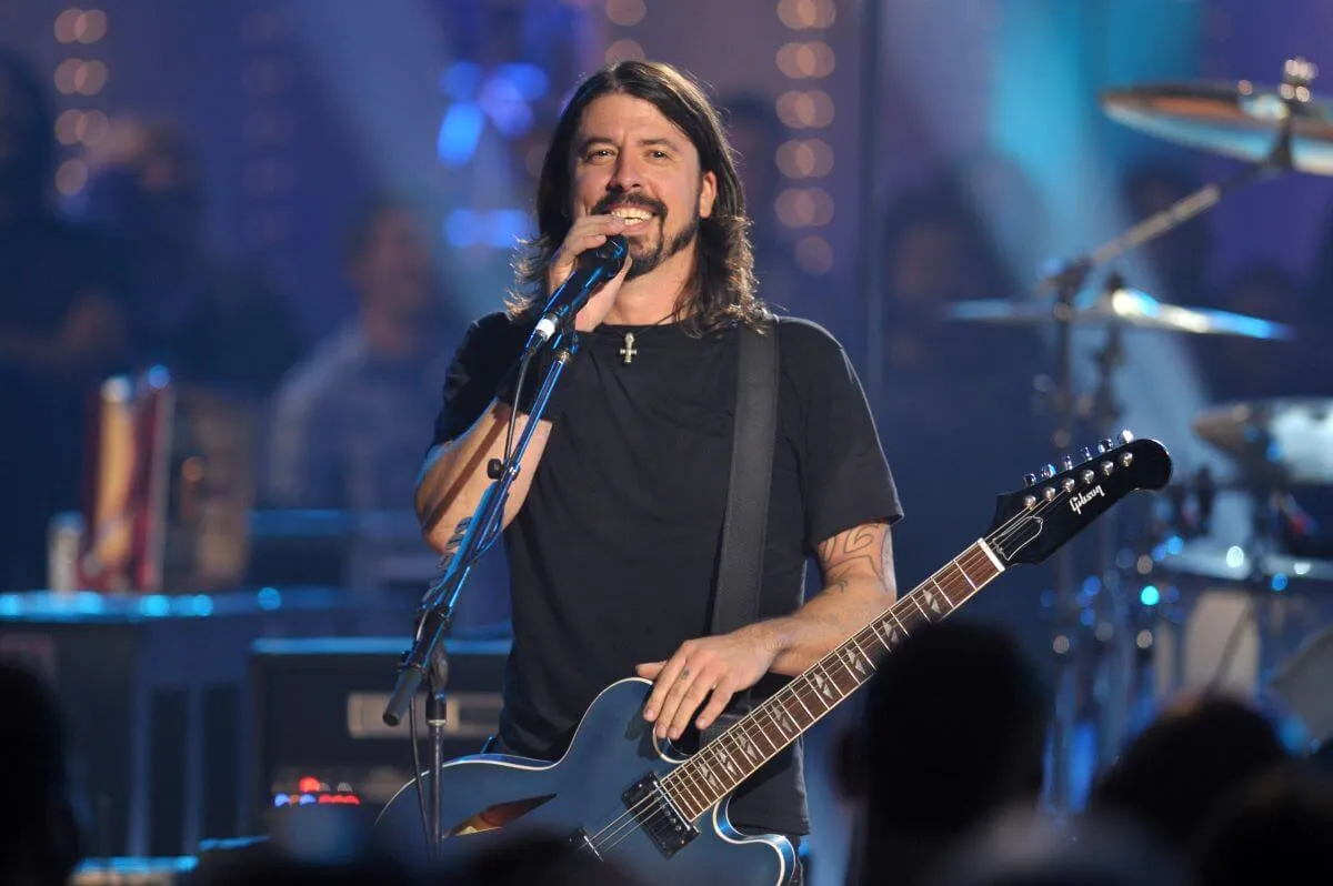 Dave Grohl wears a black shirt and talks into a microphone. He has an electric guitar.