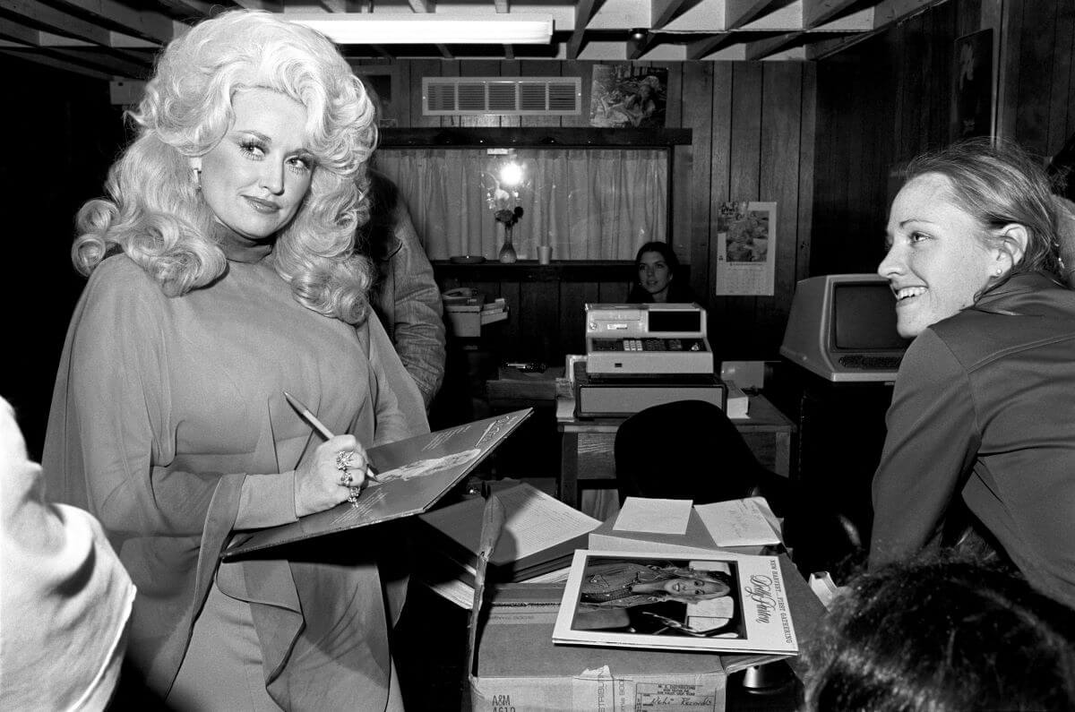 A black and white picture of Dolly Parton standing at a counter in front of a woman and signing a record.