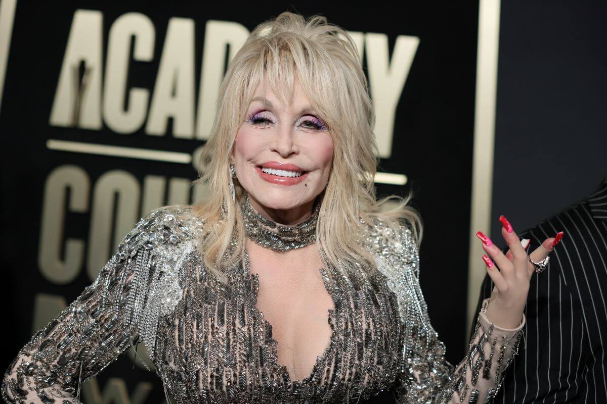 Dolly Parton wears a metallic gray dress and holds up her hand. Her nails are pink.