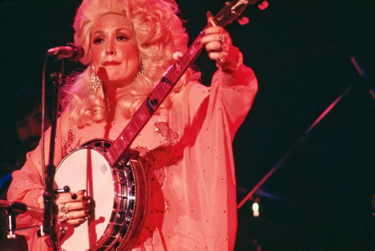 Dolly Parton stands in front of a microphone and plays the banjo.