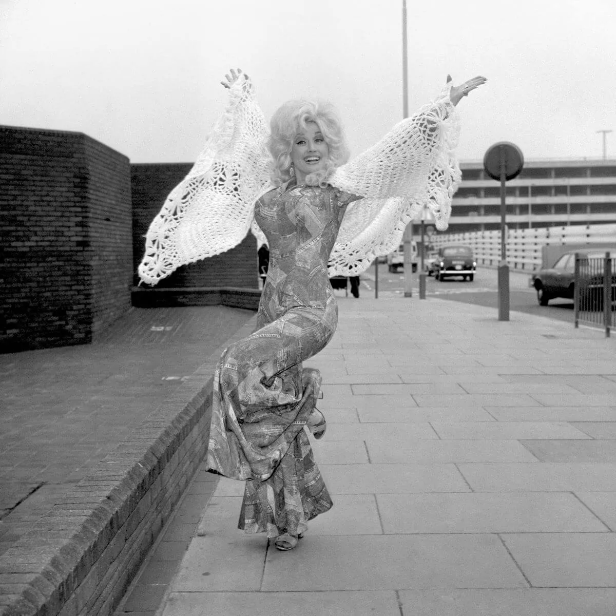 A black and white picture of Dolly Parton wearing a jumpsuit and crocheted shawl. She lifts her arms in the air and stands on one leg.