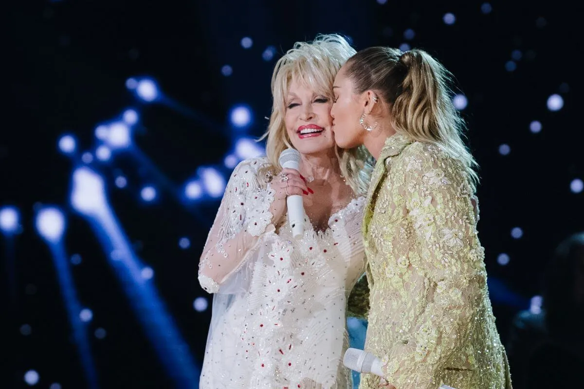 Dolly Parton wears white and holds a microphone. Miley Cyrus wears yellow and kisses Parton on the cheek.